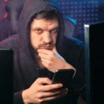 Mężczyzna w czarnej bluzie analizuje smartfon na tle cyfrowych ekranów