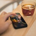 Dłoń odblokowująca smartfon obok filiżanki herbaty na drewnianym stole